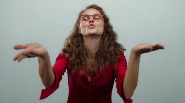 Kırmızı bluzlu, gözlüklü, öpüşen bir kadın izole edilmiş beyaz bir arka plan duvarına yaslanmış neşeli bir ifade ve vücut dili sergiliyor..