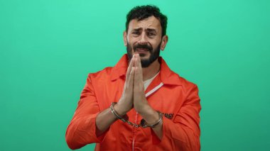 Turuncu tulumlu, sakallı ve kelepçeli, yeşil arka planda duran İspanyol adam duygusal bir ifade sergiliyor..