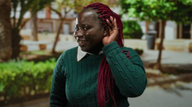 Kulaktan kulağa dikkatle dinleyen, yeşil süveter giyen, ağaçlarla çevrili ve şehir dışında yaşayan, canlı kırmızı örgüler sergileyen bir kadın..