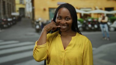 Sarı gömlekli kadın gülümsüyor ve şehir arka planında bulanık bir sokakta telefon jesti yapıyor..