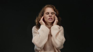 Gözlük ve süveter takan ve vücut dili ile rahatsızlık veren bir kadın, izole edilmiş siyah arka planda gürültüyü engellemek için kulaklarını kapatır.