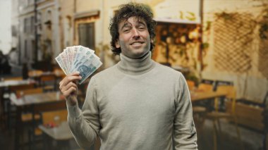 Açık hava restoranının arka planına sahip bir terasta, parlak bir şehir ortamında finansal başarı ve keyif sunan İspanyol bir adam zloti banknotları tutuyor..