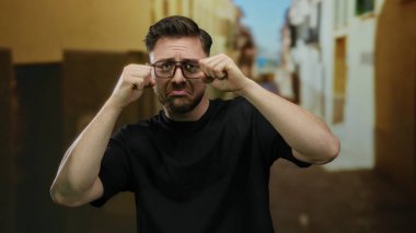 Sakallı ve gözlüklü genç bir adam, şehir sokaklarında ağlıyormuş gibi yapıyor oyuncu ifadeler ve komik duygular sergiliyor..