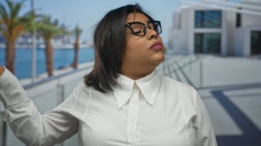 Deniz kenarındaki gezinti alanında el kol hareketi yapan genç Latin bir kadın, gözlük ve beyaz gömlek takıyor, palmiye ağaçları ve binaların arasında tavır sergiliyor..