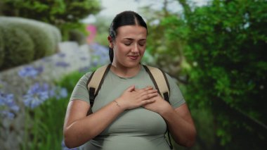 Yeşil manzaralı bir parkta, açık mavi gökyüzünün altında sırt çantalı, sıradan yeşil bir gömlek giyen bir kadın, sandığı tutmaktan rahatsızlık duyuyor..