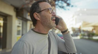 Güneşli bir şehir sokağında akıllı telefondan konuşan gözlüklü olgun bir adam, şehir ortamında modern bir yaşam tarzı ve iletişimi yansıtıyor..