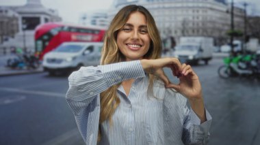 Şehrin işlek sokaklarında parmaklarıyla kalp şeklini oluştururken gülümseyen sarışın genç kadın..
