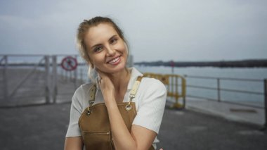 Deniz kıyısında kayıklarla önlük giymiş bulutlu bir günde metal bir çitin arkasında gülümseyen bir kadın, huzurlu denizcilik ortamında mutluluğu vurguluyor..