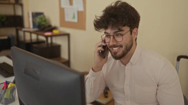 Ofis ortamında gözlüklü ve sakallı genç adam telefonda konuşuyor. Masa, bilgisayar ve raflarla çevrili, iş yerinde profesyonel iletişimle meşgul..