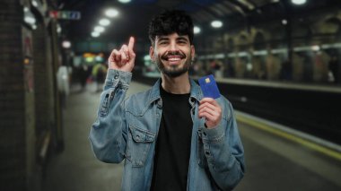 Bir tren istasyonunda elinde kredi kartı olan genç bir adam düşünceli bir görünüm sergiliyor. Finansal planlama ya da hareketli bir şehir ortamında karar verme mekanizmasını öneriyor..
