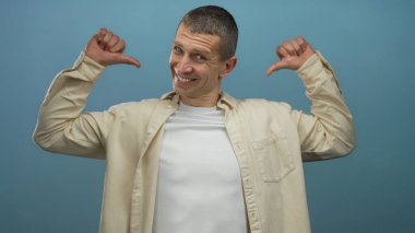 Kendine güvenen beyaz bir adam mavi izole bir arka plana gülümsüyor, bej bir gömlek ve beyaz bir tişört giyiyor, pozitif bir tavır sergiliyor..