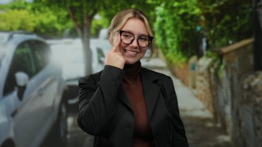 Canlı bir şehir caddesinde gözlük takan ve açık havada, yemyeşil ağaçlarla kaplı arka planda siyah bir ceket giyen bir kadın gözlerini işaret ediyor..