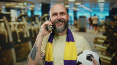 Sakallı ve dövmeli bir adam spor salonunda telefonla konuşuyor, eşarp takıyor, canlı ama rahat bir his yaratıyor modern spor aletlerinin arasında parlak ışık altında..