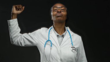 Beyaz önlüklü, steteskoplu bir kadın güçlü ve profesyonelliğini gösteren koyu arkaplana karşı yumruğunu kaldırıyor.