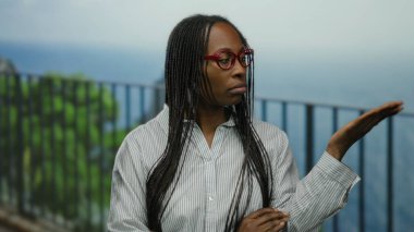 Saç örgülü bir kadın, mavi gökyüzünün altında, okyanus manzaralı ve yeşil yapraklı, düşünceli bir şekilde deniz kenarında geziniyor..