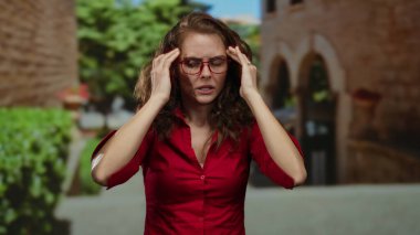 Kırmızı tişörtlü kadın şehir merkezinde hayal kırıklığını gösteriyor. Elleri başında ve gözlüklü, bulanık yeşillik ve mimariyle çevrili..