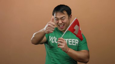 Asyalı genç bir adam elinde İsviçre bayrağı tutarak izole edilmiş kahverengi duvarın önünde gönüllü gömleği giyip baş parmak hareketleriyle heyecanını dile getiriyor.