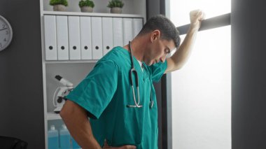 Önlüklü genç erkek sağlık çalışanı, modern klinikte cam kenarında dikiliyor. Steteskop görünür tıbbi çevreye sahip.