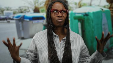 Kıvırcık saçlı bir kadın geri dönüşüm kutularının yanında dikiliyor, kollarını kavuştururken hoşnutsuzluğunu dile getiriyor, kırmızı gözlük takıyor ve sokakta çizgili bir gömlek giyiyor..