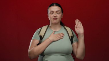 Sırt çantalı genç bir kadın kırmızı duvarın önünde yemin ediyor, eli kalbi ve gözleri kapalı, içtenlik ve bağlılık duygularını iletiyor..