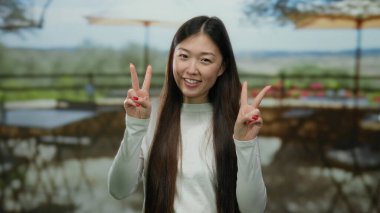 Şehir terasında gülümseyen bir kadın, uzun saçlarını ve barış jestini sergiliyor şehir restoranının arka planında, güneşli bir günde canlı, neşeli bir enerjiyi somutlaştırıyor..