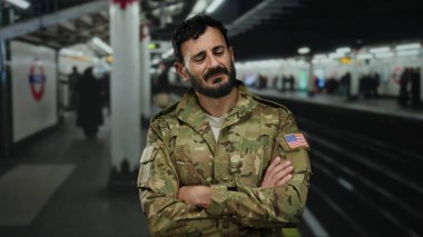 Askeri üniformalı bir adam, bir yeraltı istasyonunda kollarını kavuşturmuş, bulanık banliyö ve metro işaretleri arasında düşünceli bir yüz ifadesi sergiliyor, kentsel geçiş modunu vurguluyor..