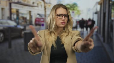 Kadın, şehir merkezinde gözlük ve bej ceket giyerken, açık havada şehir ortamını onaylamadığını belirtirken, hareketli şehir caddesinde parmaklarıyla hareket etmez..