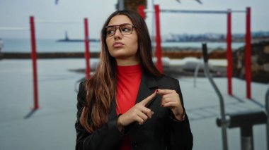 Gözlük takan bir kadın, kırmızı ekipman ve uzak kıyı şeridi olan açık hava egzersiz alanında, canlı kırmızı ve siyah giysiler içinde düşünceli bir şekilde el kol hareketi yapıyor..