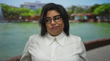 Düşünceli bir ifadeyle gözlük takan bir kadın, yemyeşil ve arka planda bir göleti olan güneşli bir parkta duruyor.