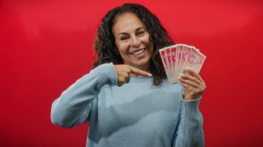 Kırmızı arka planda orta yaşlı bir kadın kendinden emin bir gülümseme ve jestle İsrail şekellerini işaret ediyor..