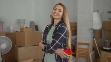 Yeni evinde kendine güvenen bir kadın etrafı kutularla ve paketlenmemiş eşyalarla çevrili, akıllı telefon elinde, parlak bir iç mekanda yeni bir başlangıcın heyecanını simgeliyor..