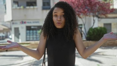 Güneşli bir sokakta, şehir binaları ve ağaç gölgeleriyle çevrili, aydınlık bir sokakta dikilen genç kadın bir şaşkınlık ve merak anını yakalıyor..