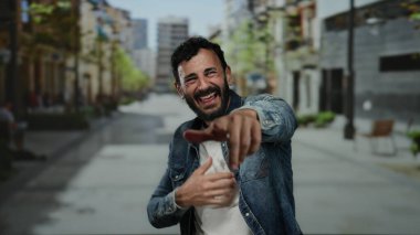 Sakallı İspanyol bir adam gülerek ve bir şehir sokağında, kot ceket giyip dışarıda mutluluğu ifade ederek neşeyle işaret ediyor..