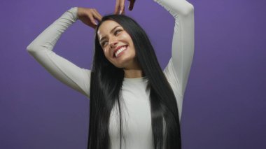 Genç İspanyol kadın, elleri mor arka planda bir kalp şekli oluşturur, izole edilmiş bir ortamda neşeyle gülümser..