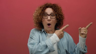 Orta yaşlı İspanyol bir kadın şok edici bir ifadeyi gösteren canlı kırmızı bir arkaplan üzerinde gözlüklü ve kot gömlekli birini işaret ederken şaşırdı..