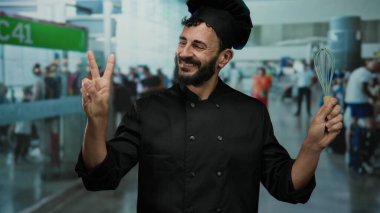 Sakallı İspanyol aşçı elinde çırpıcı ve havaalanı terminalinde gülümsüyor, neşeli bir seyahat ortamı yaratıyor..