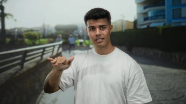 Gündelik beyaz tişörtlü genç adam modern binalar ve arka planda yeşillik olan bir şehir caddesinde güvenle duruyor, kentsel yaşam tarzı ve kendine güveni aktarıyor..