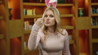 Kitaplıklarla çevrili bir kütüphanede komik bir şekilde duran tavşan kulaklı kadın, neşeli bir ifadeyi açık yüreklilikle ifade ediyor..