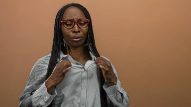 Gözlüklü kadın gömlek yakasını kahverengi izole bir arka plana göre ayarlıyor. Düşünceli bir ifade ve uzun kıvırcık saçla ısı rahatsızlığını gösteriyor..