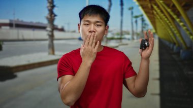 Şehrin caddesinde kırmızı gömlekli genç adam araba anahtarlarını tutuyor. Arka planda el hareketi, palmiye ağacı ve kentsel çevre ile şaşkınlığını dile getiriyor..