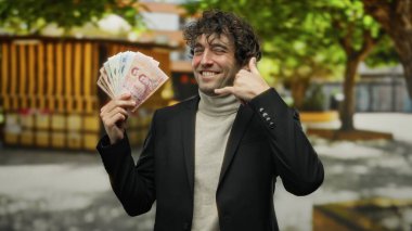 Hispanik bir adam şehrin dışında, Thai banknotlarını tutarken bir telefon jesti yapıyor, canlı bir şehir ortamında finansal başarı ve iletişimi öneriyor..