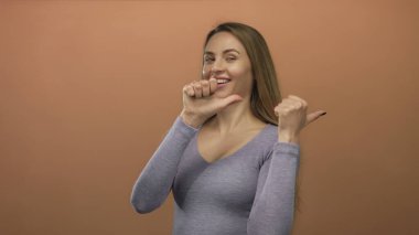 İzole edilmiş kahverengi duvarın üzerinden gülümseyen bir kadın iki başparmağını da kendine güvenle gösteren gri bir gömlek giyen mutluluğu ve kendine güveni gösteren bir kadın..