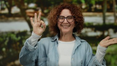 Parkta gülümseyen, kıvırcık saçlı ve gözlüklü, kot gömlek giyen, yeşil arka planda bulanık bir kadın..