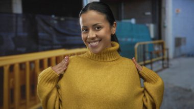 İnşaat alanında sarı bir kazakla gülümseyen bir kadın, açık havada, şehir ortamında mutluluk ve güven yayıyor..