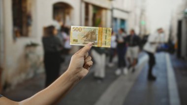 Şehrin işlek caddesinde Norveç kronu banknotları tutan adam, arka planda bulanık yayalar ve tarihi mimariyle canlı şehir manzarası sergiliyor..