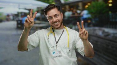 Genç bir adam, sakallı ve steteskoplu beyaz bir doktor şehir caddesinde zafer işaretleri yapıyor, hareketli bir şehir ortamında kameraya güvenle gülümsüyor..