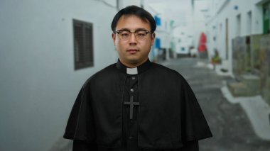 Şehir ortamında sakin dar bir sokakta duran Asyalı bir rahip, gözlük takıyor ve haç kolyeli siyah bir rahip cübbesi takıyor, sakin ve inançlı bir şekilde.