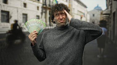 Rublemleri olan bir adam şehrin dışında sıkıntılı görünüyor, elinde para tutuyor, süveter giyiyor, gündüz vakti, yüzünde karışık duygular, güneşli bir hava..