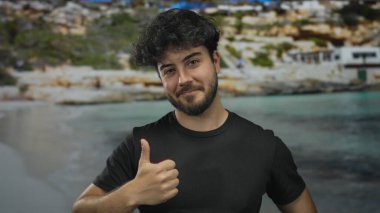 Genç sakallı bir adam, berrak mavi bir deniz ve kayalık bir kıyı şeridi ile manzaralı bir plaj zemininde olumlu ve neşeli bir hava yayıyor..