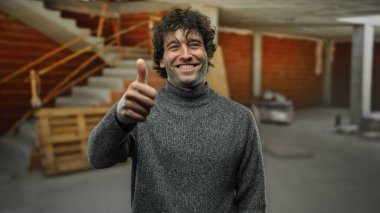 İnşaat alanında parmakları havada, gri süveter giymiş, bitmemiş duvarlar ve tahta kirişlerle çevrili, olumlu ve onaylı bir şekilde gülümseyen genç adam..